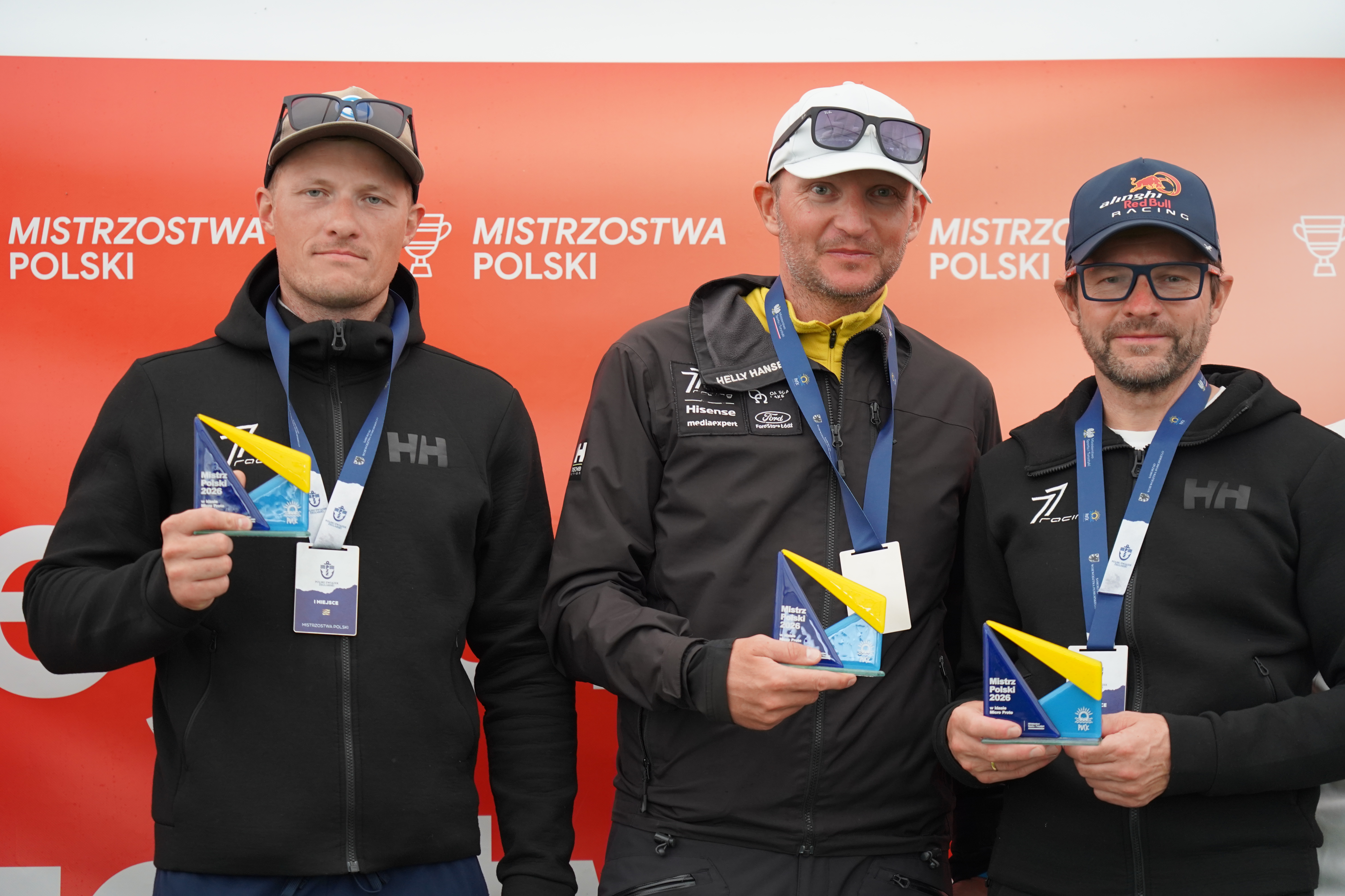 Trzej żeglarze w bluzach 77 Racing trzymają statuetki przed ścianką z napisem Mistrzostwa Polski