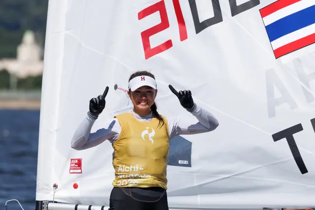 Sophia Montgomery w złotej koszulce Aichi-Nagoya 2026 unosi palce wskazujące na tle żagla ILCA z flagą Tajlandii
