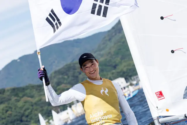 Uśmiechnięty Jeemin Ha w złotej koszulce unosi flagę Korei Południowej na łódce ILCA 7 w Gamagori