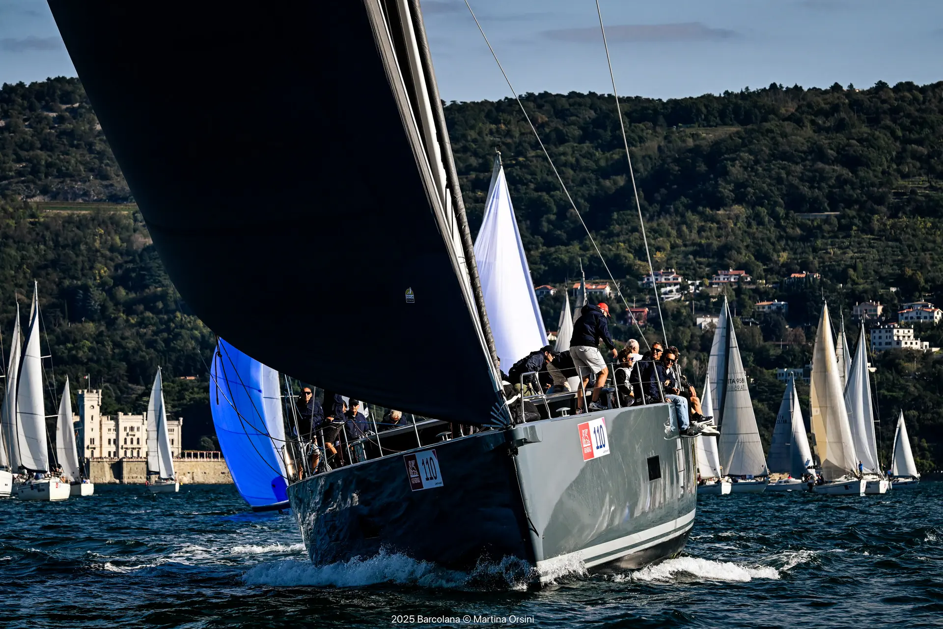 Jacht z numerem 110 pod czarnym grotem na tle floty i zalesionego wybrzeża z białym zamkiem nad wodą podczas Barcolana57