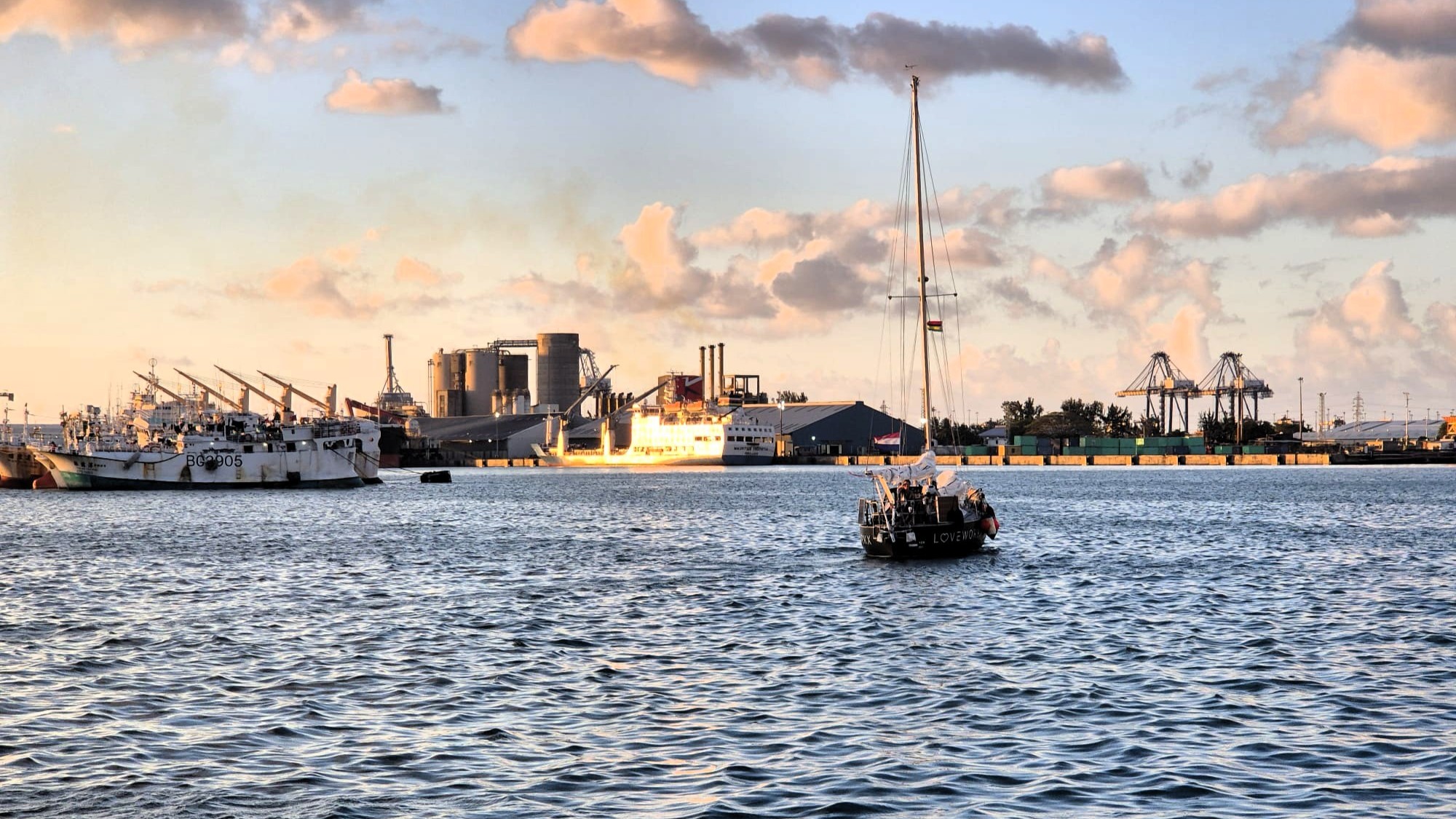 Czarny jacht żaglowy LOVEWORKX na wodzie portu Port Louis w złotym świetle, w tle dźwigi i statki