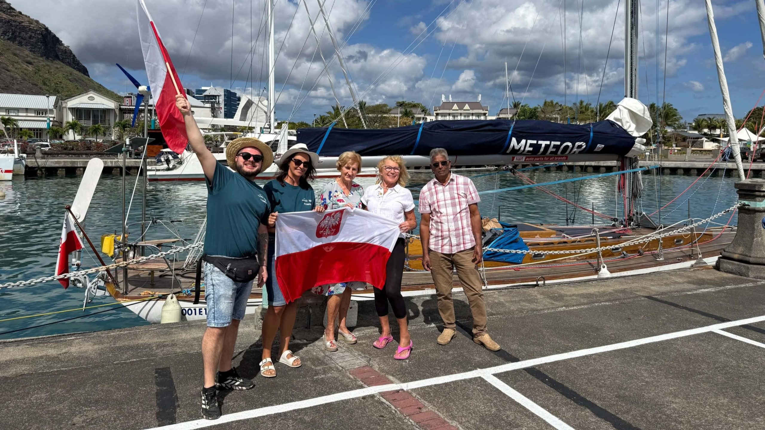 Pięć osób z biało-czerwoną flagą przy jachcie Meteor zacumowanym w Port Louis