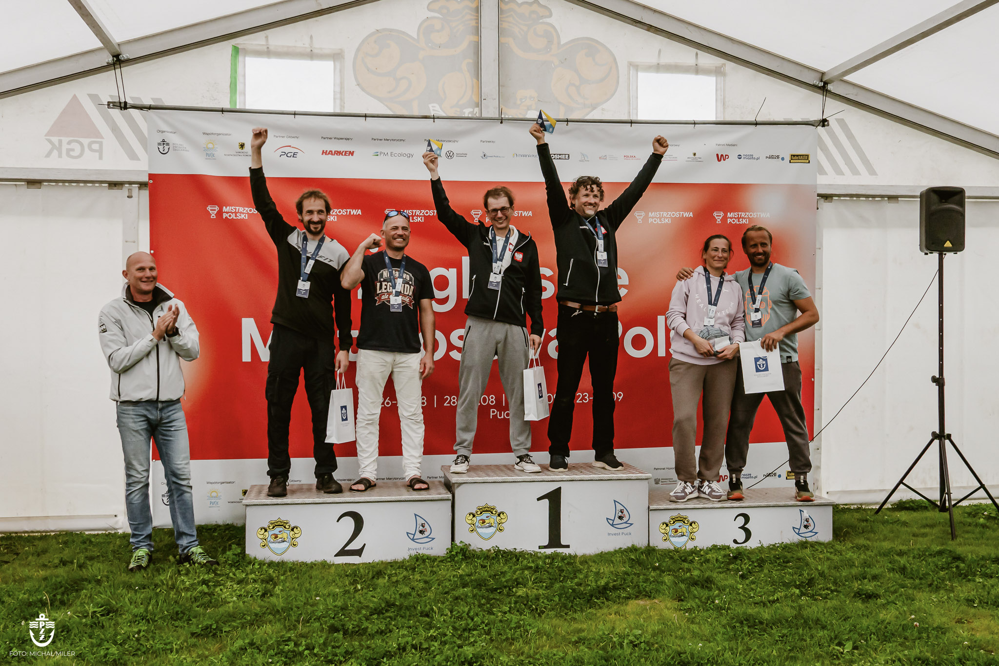 Załogi z uniesionymi rękami na trzystopniowym podium pod namiotem