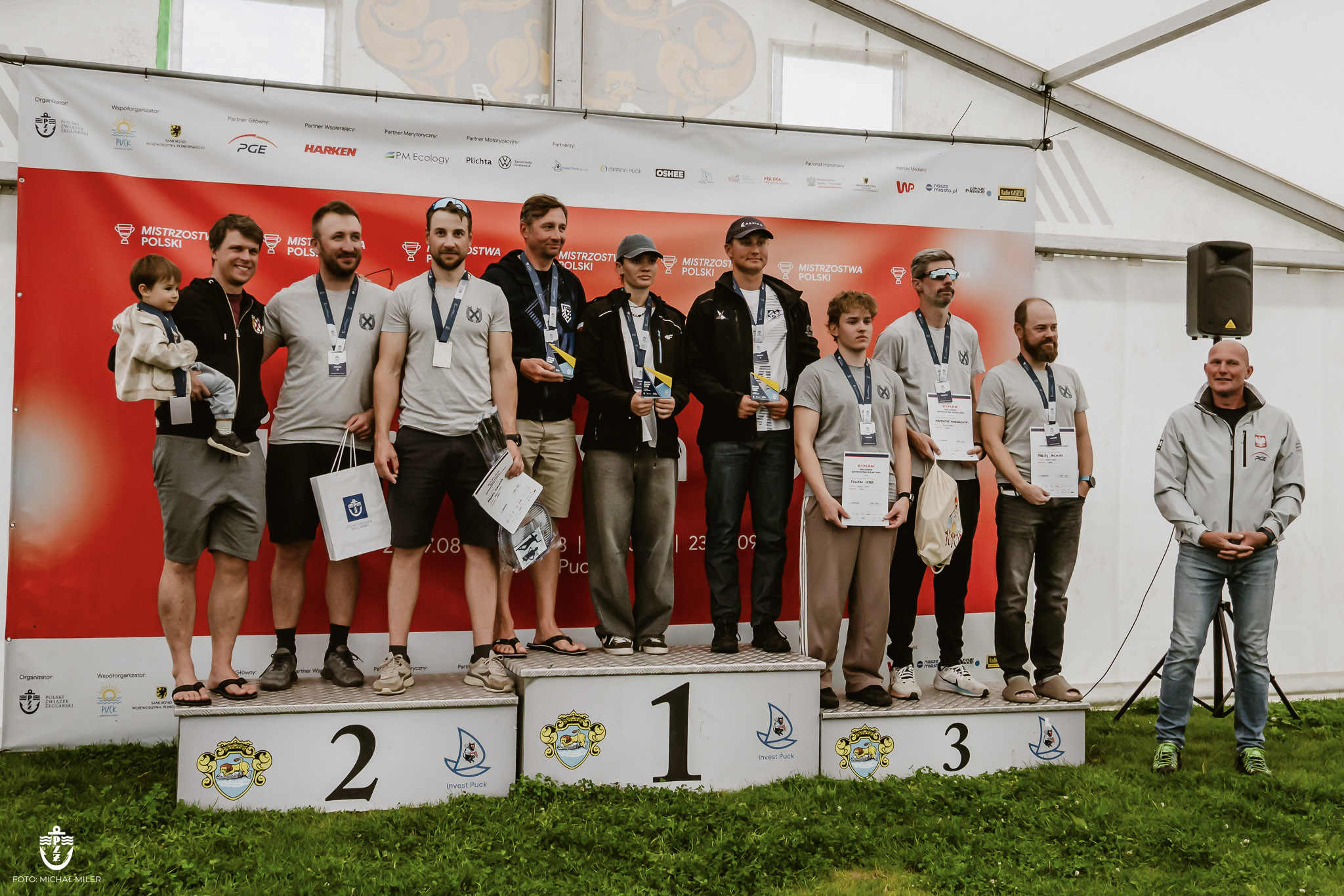 Medaliści z dyplomami stoją na trzystopniowym podium przed ścianką sponsorów