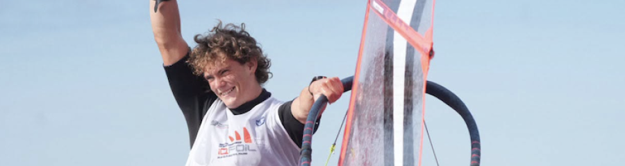 Uśmiechnięty windsurfer w koszulce MŚ U23 iQFOiL unosi rękę obok żagla