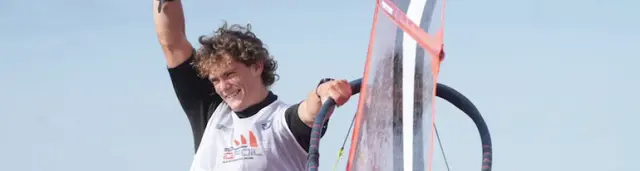 Uśmiechnięty windsurfer w koszulce MŚ U23 iQFOiL unosi rękę obok żagla