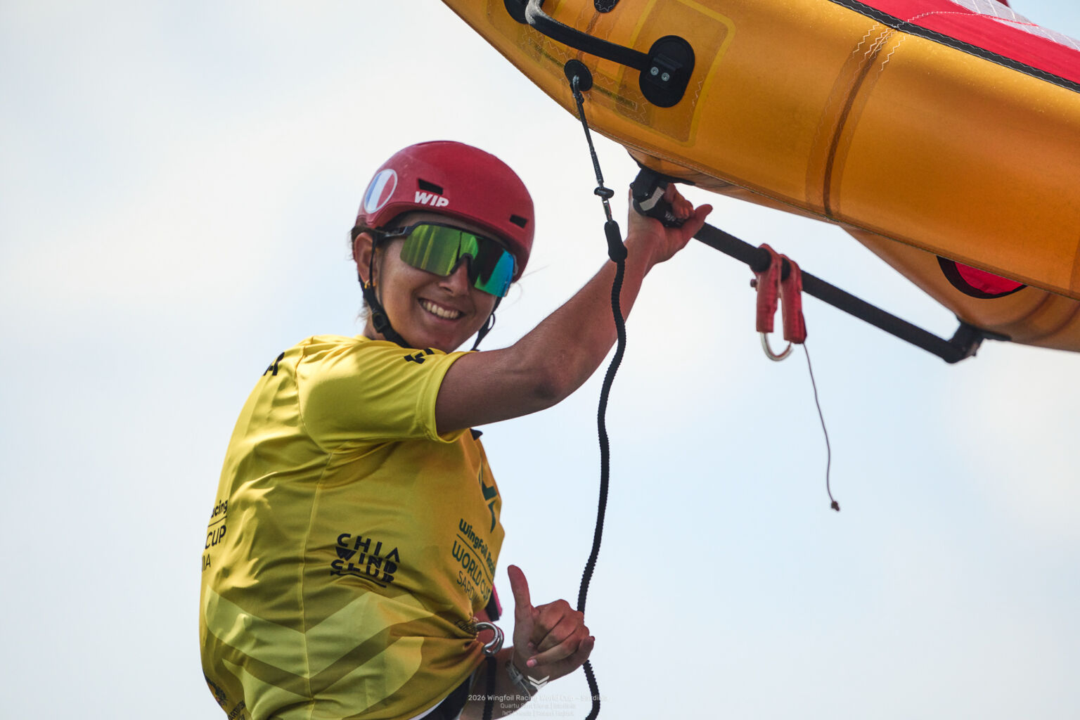 Zawodniczka wingfoil w czerwonym kasku i żółtej koszulce trzyma skrzydło podczas Pucharu Świata na Sardynii